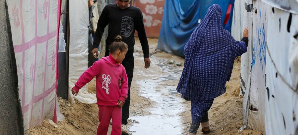 امرأة ترتدي حجاباً أزرق تسير عبر طريق موحل في مخيم خيام في غزة، برفقة رجل وفتاة صغيرة، بينما ينظر إليها شخص آخر.