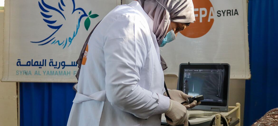 A female healthcare worker in a white coat and hijab performs a medical examination using a tablet device at a UNFPA mobile health clinic in Syria, supporting displaced women and girls with reproductive health services.