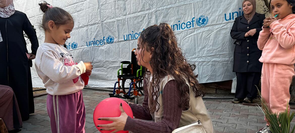 A young girl in Gaza, supported by UNICEF, plays with a red ball while a UNICEF worker kneels beside her. The scene highlights UNICEF's efforts to provide rehabilitation, mental health support, and inclusive education to children with disabilities in Gaza.