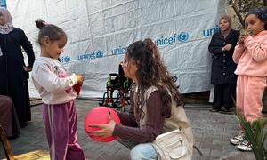 A young girl in Gaza, supported by UNICEF, plays with a red ball while a UNICEF worker kneels beside her. The scene highlights UNICEF's efforts to provide rehabilitation, mental health support, and inclusive education to children with disabilities in Gaza.