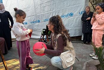 A young girl in Gaza, supported by UNICEF, plays with a red ball while a UNICEF worker kneels beside her. The scene highlights UNICEF's efforts to provide rehabilitation, mental health support, and inclusive education to children with disabilities in Gaza.