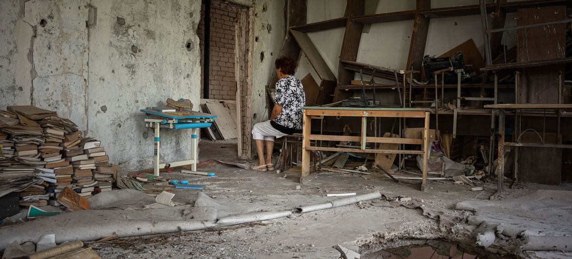 A woman, identified as Maryna, sits in a damaged school in war-torn Ukraine, surrounded by debris and broken furniture, reflecting on her past as a teacher before the invasion.