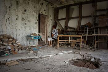A woman, identified as Maryna, sits in a damaged school in war-torn Ukraine, surrounded by debris and broken furniture, reflecting on her past as a teacher before the invasion.