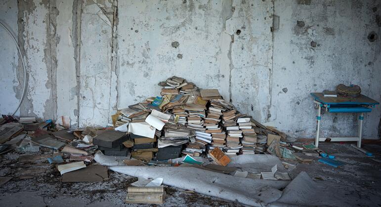 Поврежденная школа в Украине с кучами книг и разбитым столом, символизируя влияние войны и сексуального насилия на образование и повседневную жизнь.