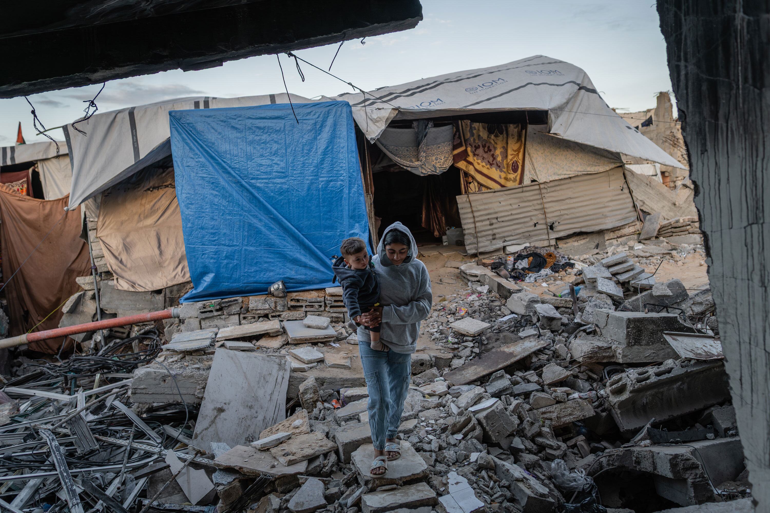 A woman, Sama, holds her 10-month-old child, Akram, as they stand amidst the rubble of their destroyed home in Al-Zarqaa, Gaza City. The family lives in a makeshift tent and relies on WFP assistance and hot meals from humanitarian organizations.