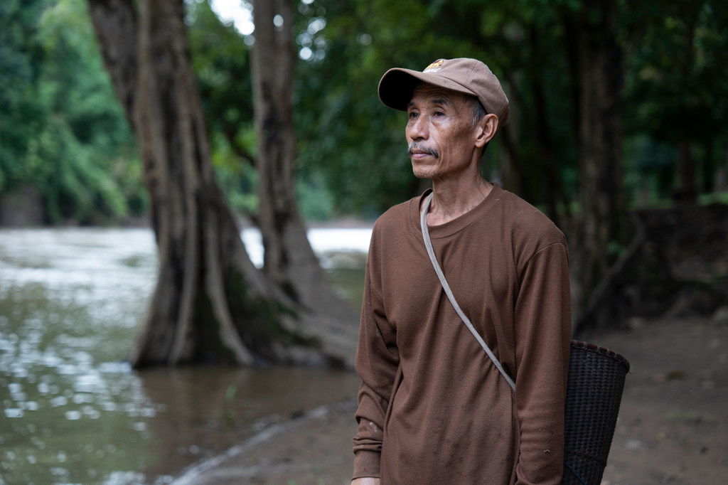 An elderly Indigenous Karen man from Thailand stands by a river in a forested landscape.