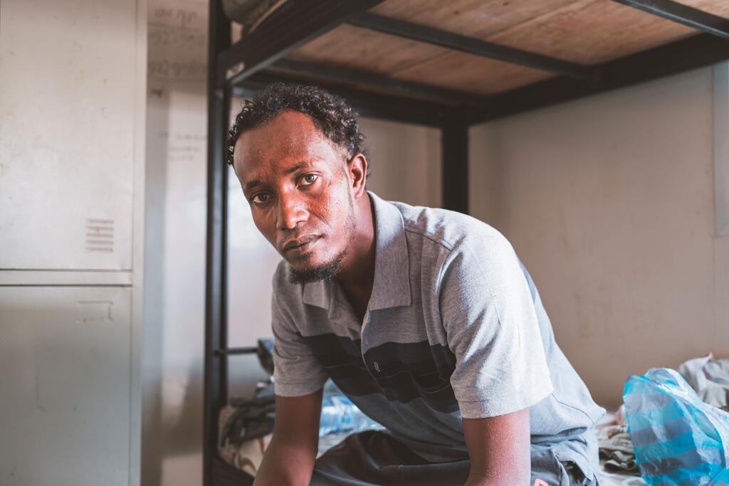 Portrait of Jamal, an Ethiopian migrant in his early thirties, sitting on a bunk bed inside a shelter in Djibouti. He is looking directly at the camera with a serious expression, wearing a gray and black striped polo shirt.