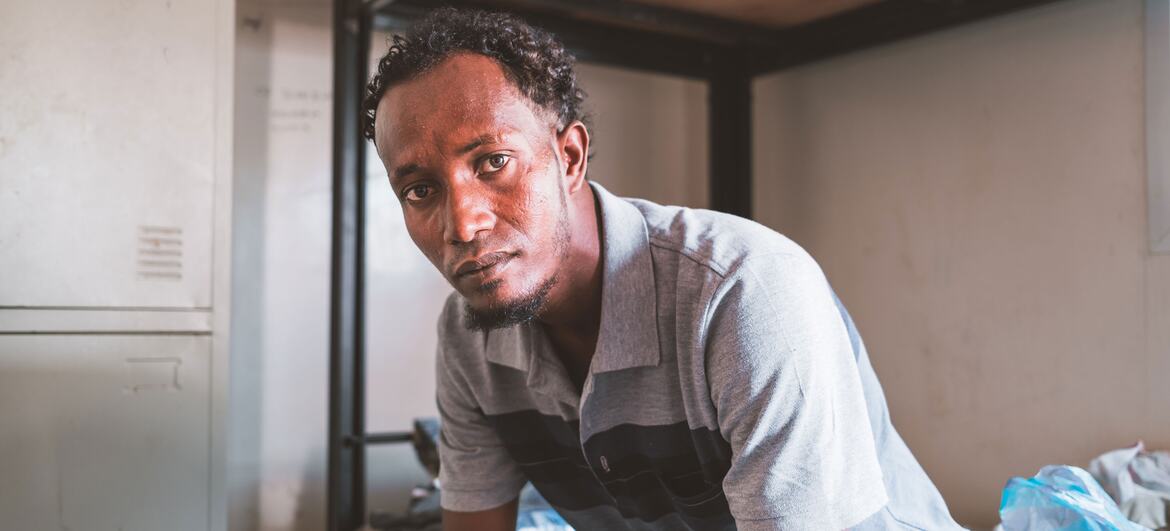 Portrait de Jamal, un migrant éthiopien d'une trentaine d'années, assis sur un lit superposé à l'intérieur d'un refuge à Djibouti. Il regarde directement la caméra avec une expression sérieuse, portant un polo à rayures grises et noires.