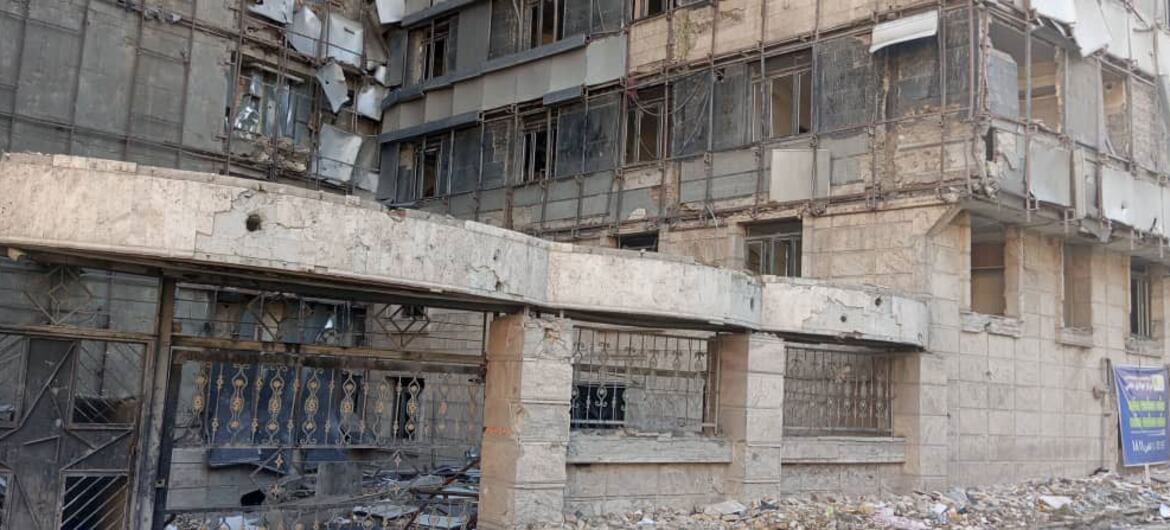A low-angle photograph of a heavily damaged multi-story concrete building in Tehran, Iran. The facade shows extensive destruction with exposed concrete, broken windows, and scattered rubble on the ground. Scaffolding is visible on the upper floors.