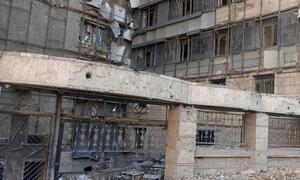 A low-angle photograph of a heavily damaged multi-story concrete building in Tehran, Iran. The facade shows extensive destruction with exposed concrete, broken windows, and scattered rubble on the ground. Scaffolding is visible on the upper floors.