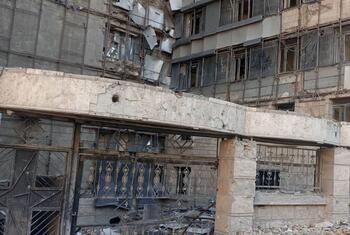 A low-angle photograph of a heavily damaged multi-story concrete building in Tehran, Iran. The facade shows extensive destruction with exposed concrete, broken windows, and scattered rubble on the ground. Scaffolding is visible on the upper floors.