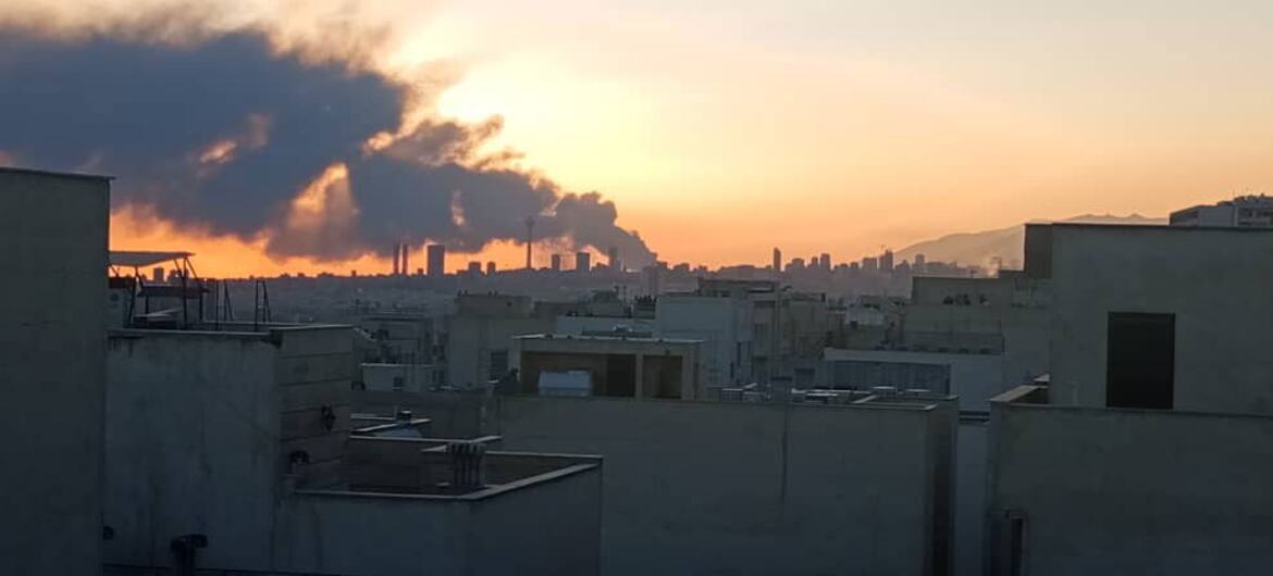 منظر بانورامي على أفق طهران عند غروب الشمس، مع نغمة كبيرة من الدخان الداكن ترتفع في المسافة خلف المباني الخرسانية في المدينة.