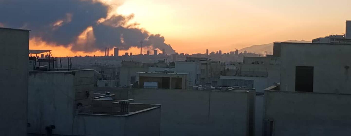 Uma vista panorâmica do horizonte de Teerã ao pôr do sol, com uma grande pluma de fumaça escura subindo na distância atrás dos edifícios de concreto da cidade.