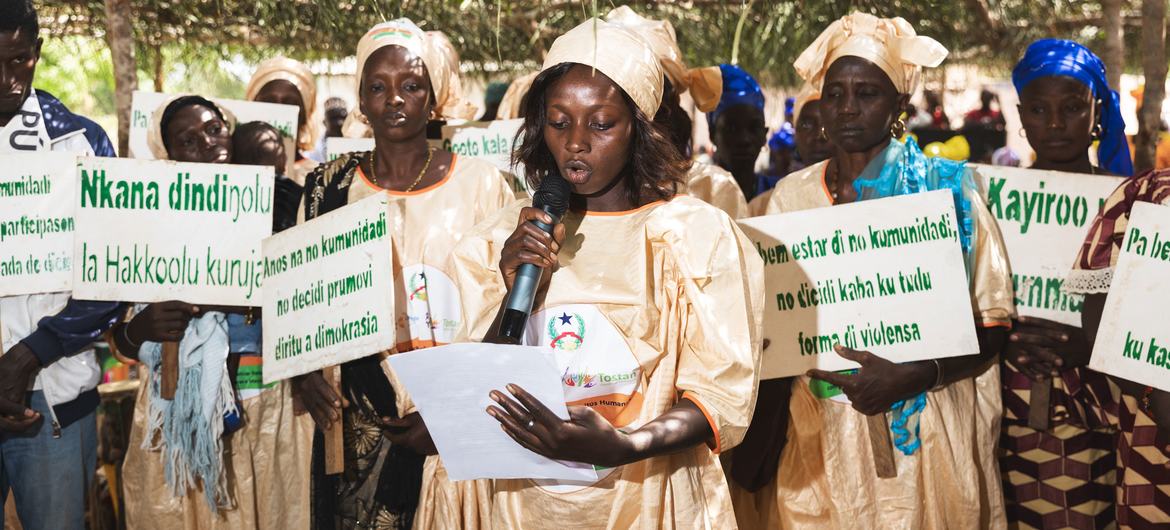 Uma mulher de vestido bege fala em um microfone em um encontro ao ar livre na Guiné-Bissau, cercada por outros membros da comunidade segurando cartazes contra a mutilação genital feminina.
