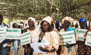 Uma mulher de vestido bege fala em um microfone em um encontro ao ar livre na Guiné-Bissau, cercada por outros membros da comunidade segurando cartazes contra a mutilação genital feminina.