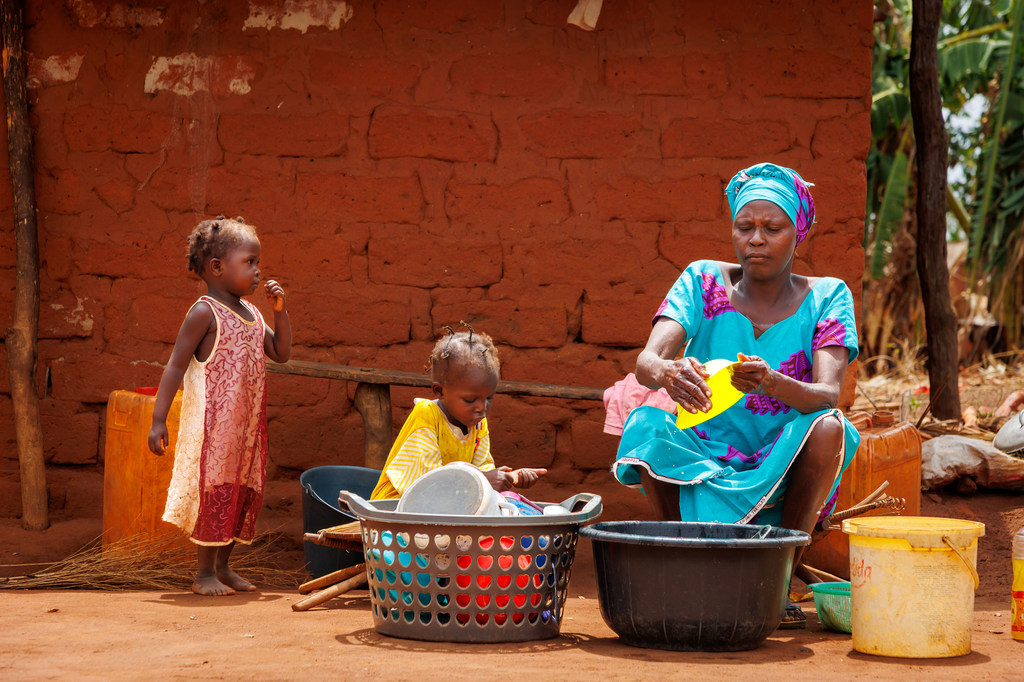 Une femme et deux jeunes enfants dans un village de la République centrafricaine ; la femme fait la vaisselle devant un bâtiment en terre rouge.