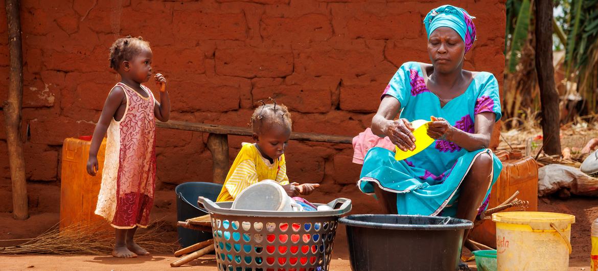 A woman and two young children in a village in the Central African Republic, with the woman washing dishes in front of a red clay building.
