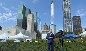 Mwanamume aliyevaa suti amesimama nje mbele ya kamera kwenye tripod, huku makao makuu UMOJA WA MATAIFA na anga ya juu ya New York vikiwa nyuma.