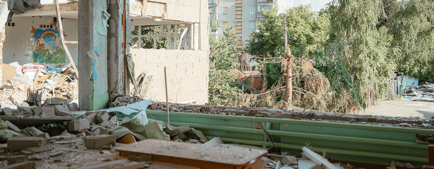 Intérieur en ruine d'une école maternelle endommagée à Tchouhouïv, en Ukraine, montrant des tables en bois renversées, des murs brisés et des décombres.