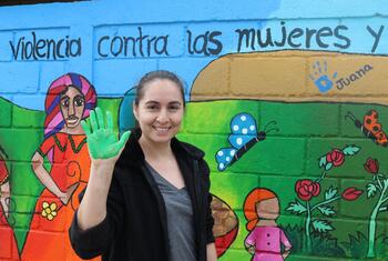 Une femme avec une main peinte en vert se tient devant une peinture murale colorée qui dit "Violencia contra las mujeres y" (Violence contre les femmes et), promouvant la prise de conscience de la violence fondée sur le genre au Honduras pendant les 16 jours d'activisme.
