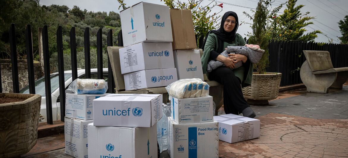 Une femme déplacée portant un hijab et tenant un bébé est assise à côté d'une grande pile de boîtes d'aide alimentaire de l'UNICEF et du PAM au Liban.