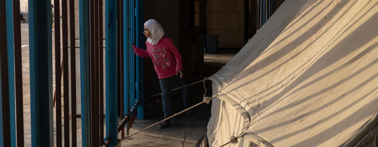 Une jeune fille vêtue d'une chemise rose et d'un hijab blanc regarde à travers une porte métallique bleue une grande tente beige dans un site d'aide du PAM au Liban.