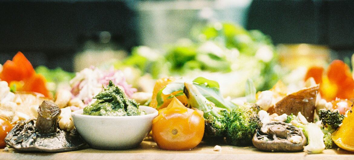 Una vista de cerca de una bandeja de madera llena de verduras sobrantes que incluyen setas, tomates y brócoli, ilustrando el desperdicio de alimentos.