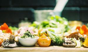 Una vista de cerca de una bandeja de madera llena de verduras sobrantes que incluyen setas, tomates y brócoli, ilustrando el desperdicio de alimentos.