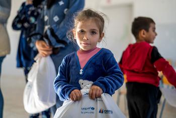 فتاة صغيرة تحمل حقيبة بيضاء عليها شعارات UNICEF وأييرا، تتلقى ملابس شتوية ومستلزمات مدرسية.