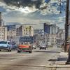 Una vibrante escena callejera en La Habana, Cuba, con coches clásicos de época conduciendo junto a edificios históricos bajo un cielo parcialmente nublado.