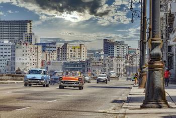 Una vibrante escena callejera en La Habana, Cuba, con coches clásicos de época conduciendo junto a edificios históricos bajo un cielo parcialmente nublado.