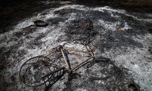 Le cadre métallique calciné d'un vélo gisant sur un sol brûlé recouvert de cendres et de débris.