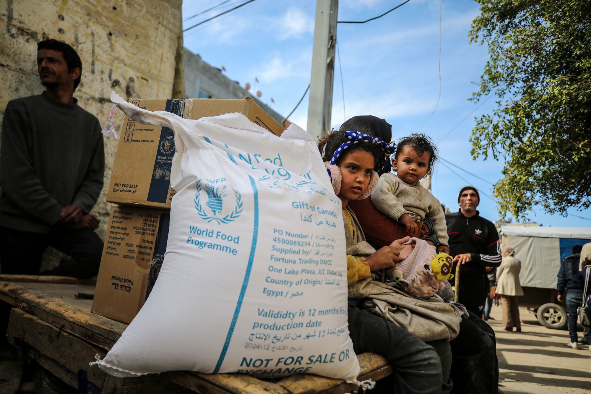 فتاة فلسطينية صغيرة تحمل طفلاً رضيعاً وهي جالسة على شاحنة خشبية بجوار كيس كبير من مساعدات برنامج الأغذية العالمي في مخيم النصيرات بقطاع غزة.
