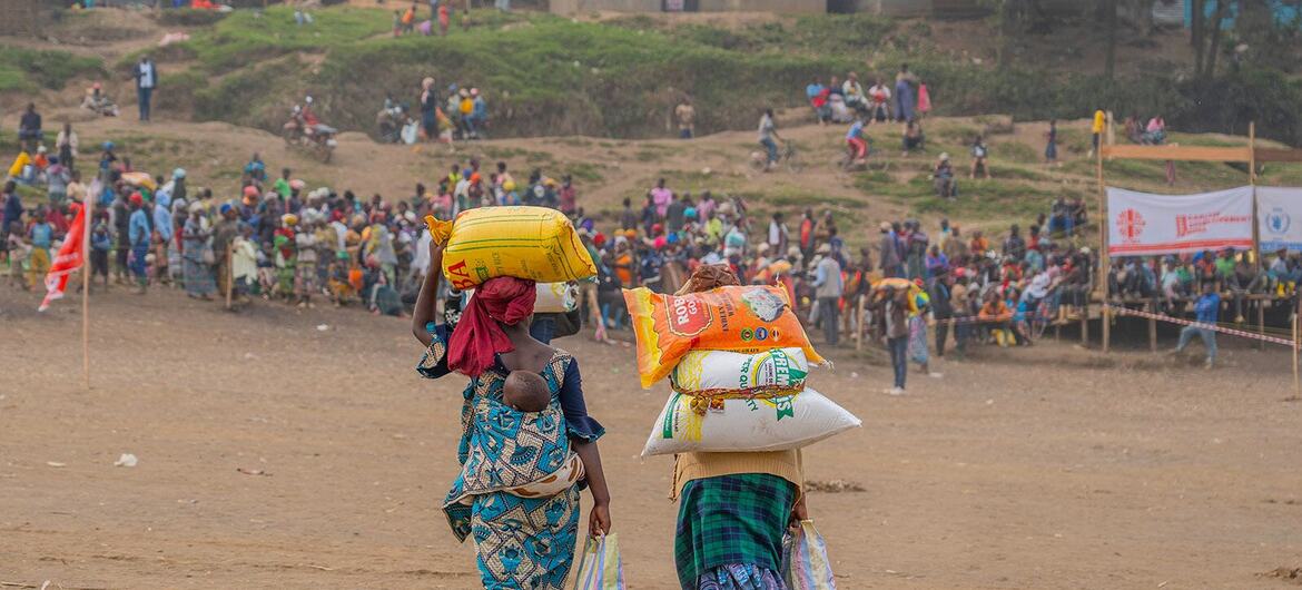 Two women carrying sacks of food items walk away from the camera in a crowded outdoor setting, likely part of a WFP food assistance program in the DRC.