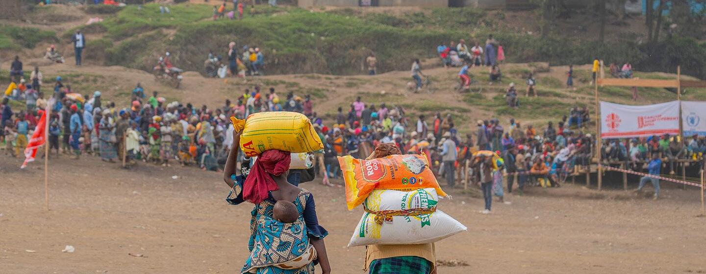 Deux femmes portant des sacs d'articles alimentaires s'éloignent de la caméra dans un cadre extérieur bondé, probablement dans le cadre d'un programme d'aide alimentaire du PAM en RDC.