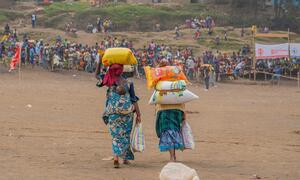 Two women carrying sacks of food items walk away from the camera in a crowded outdoor setting, likely part of a WFP food assistance program in the DRC.