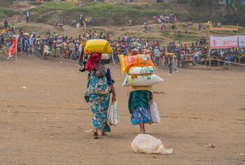 Two women carrying sacks of food items walk away from the camera in a crowded outdoor setting, likely part of a WFP food assistance program in the DRC.