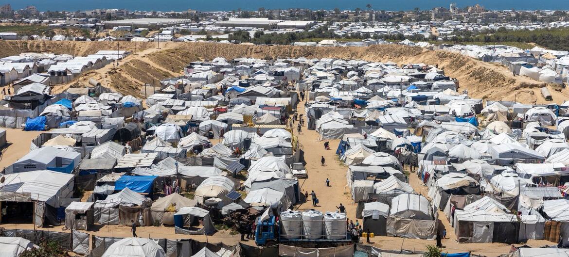 An aerial view of a large displacement camp in Gaza filled with numerous temporary tents and shelters, with a coastal city and the sea in the background.