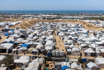 An aerial view of a large displacement camp in Gaza filled with numerous temporary tents and shelters, with a coastal city and the sea in the background.