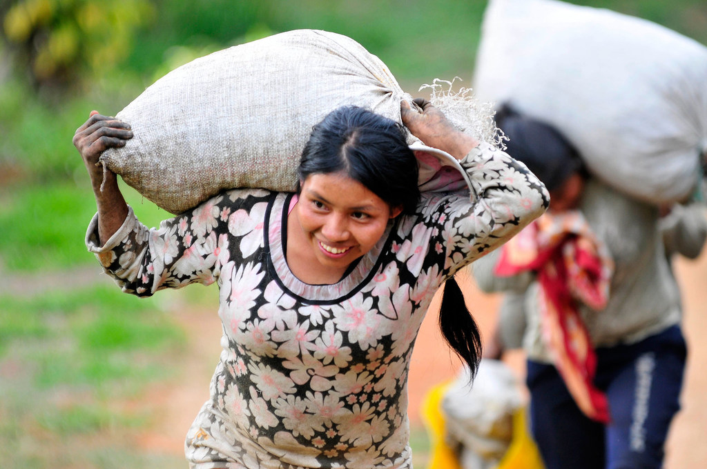 Une femme souriante, vêtue d'une chemise à fleurs, porte un grand sac sur l'épaule dans un champ rural colombien.