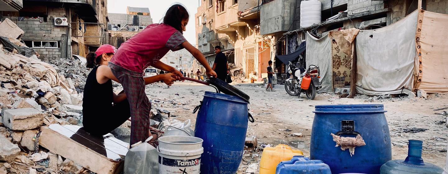 Des enfants vont chercher de l'eau dans de grands récipients, dans une rue jonchée de gravats et de débris à Gaza.