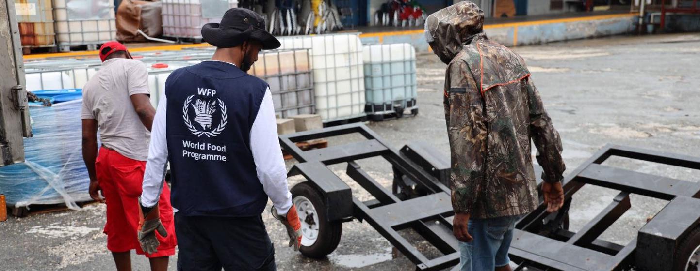Trois hommes, dont un porte un gilet du Programme alimentaire mondial, travaillent sur un quai de chargement avec une remorque et des conteneurs d'eau.