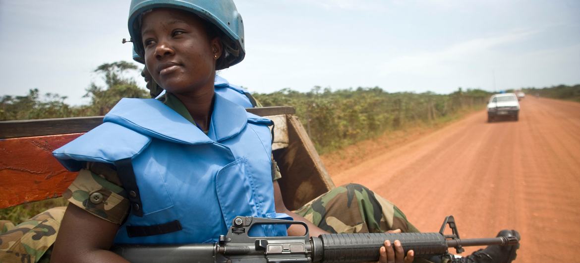 Uma integrante das forças de paz da ONU , vestindo uniforme e capacete azul, patrulha Buchanan, na Libéria, com o GhanaBat. Ela segura um fuzil de assalto enquanto está sentada em um veículo em uma estrada de terra.