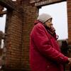 Una anciana con una chaqueta roja y un sombrero beige se encuentra junto a las ruinas de una casa de ladrillos, sosteniendo un bastón y cubriendo su boca con su mano, reflejando su vida y el impacto de la guerra en su casa en Staryi Saltiv, Ucrania.