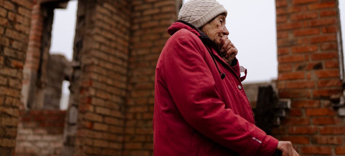 Une femme âgée en veste rouge et chapeau beige se tient à côté des ruines d'une maison en brique, tenant un bâton et couvrant sa bouche avec sa main, en réfléchissant à sa vie et à l'impact de la guerre sur sa maison à Staryi Saltiv, en Ukraine.