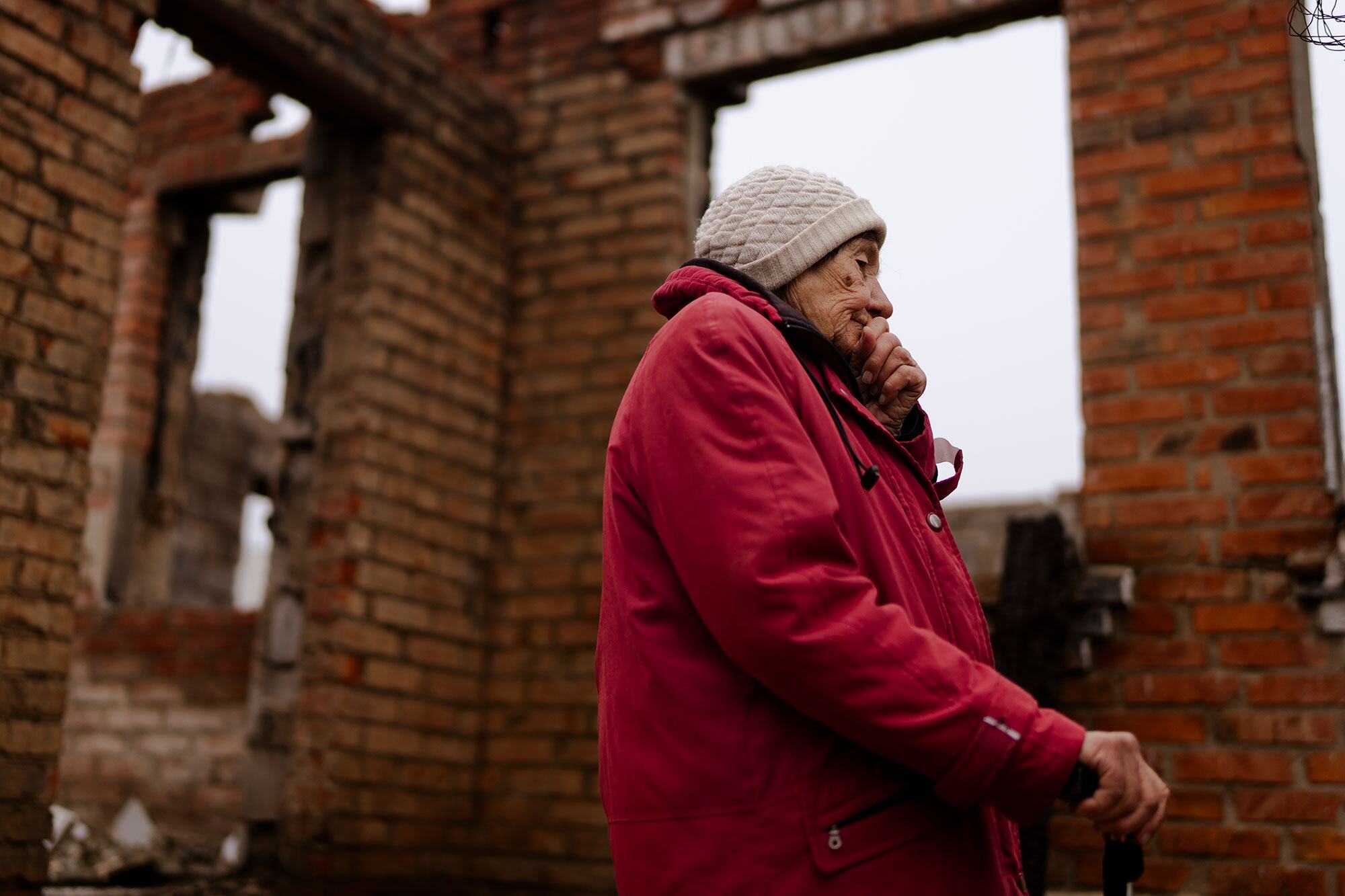 Une femme âgée en veste rouge et chapeau beige se tient à côté des ruines d'une maison en brique, tenant un bâton et couvrant sa bouche avec sa main, en réfléchissant à sa vie et à l'impact de la guerre sur sa maison à Staryi Saltiv, en Ukraine.