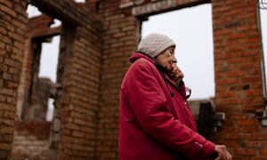 Una anciana con una chaqueta roja y un sombrero beige se encuentra junto a las ruinas de una casa de ladrillos, sosteniendo un bastón y cubriendo su boca con su mano, reflejando su vida y el impacto de la guerra en su casa en Staryi Saltiv, Ucrania.