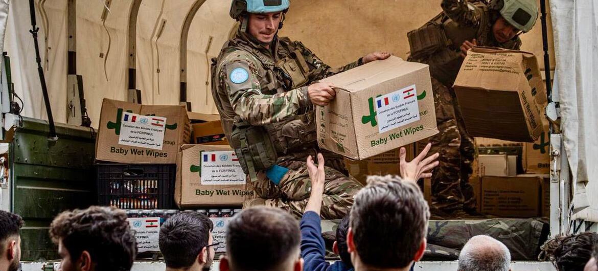 UNIFIL peacekeepers in camouflage uniforms and blue helmets unload humanitarian aid supplies, including food and medicine boxes labeled 'Baby Wet Wipes' and marked with UN and French flags, from a military truck to civilians in Lebanon.