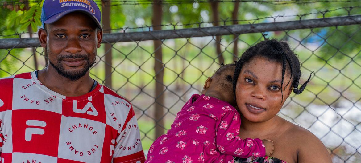 Una familia deportada permanece reunida en el centro de deportación. Un hombre con una camisa a cuadros rojos y blancos y una gorra azul está junto a una mujer que sostiene a un niño pequeño. Se encuentran frente a una cerca de alambre de púas.