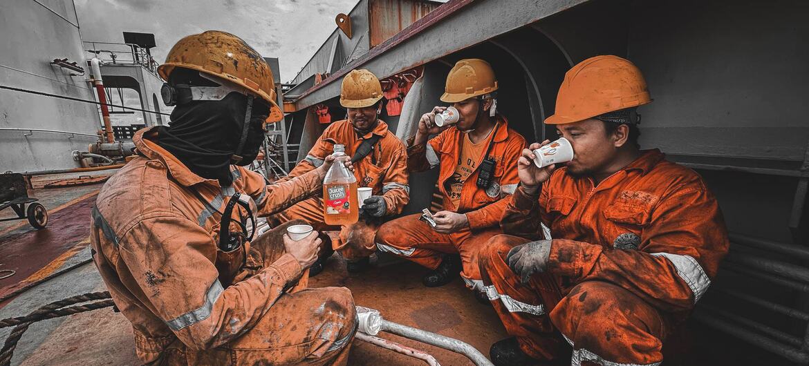Four seafarers in orange coveralls and hard hats taking a break on the deck of a ship, drinking from cups and sharing a bottle of drink.